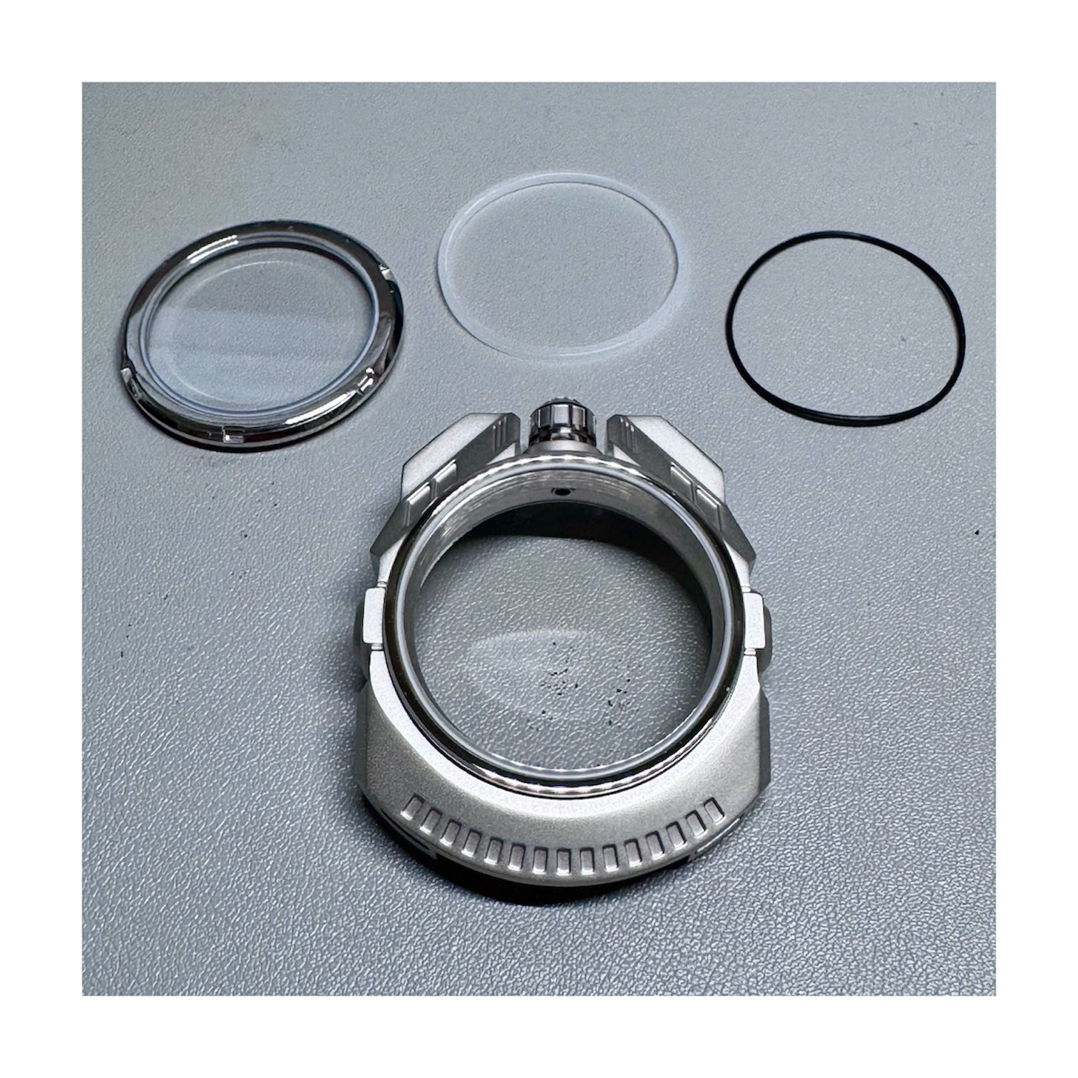 Mechanical watch components including a metal ring, glass cover, and rubber gasket on a gray background.