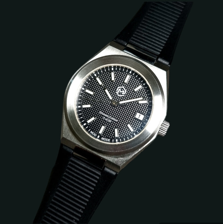 A silver-toned Project MM1 Pioneer Edition watch with a black waffle dial, stainless steel bracelet, and a vulcanised rubber strap.